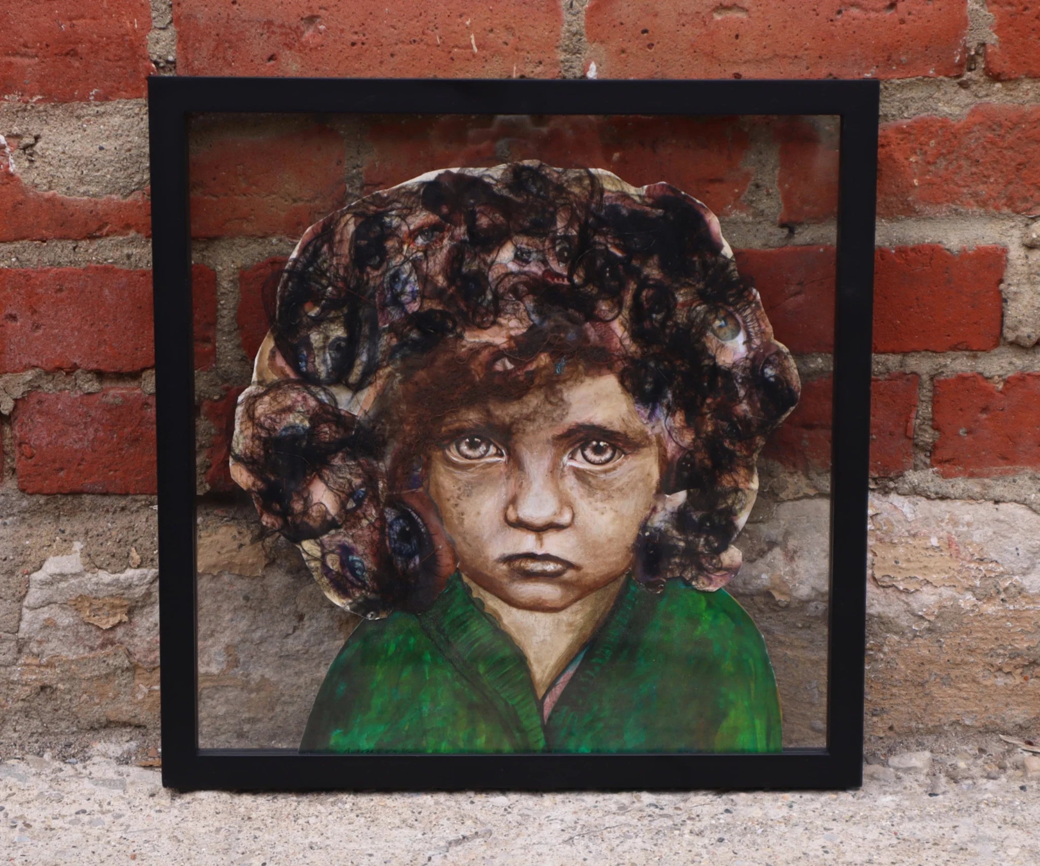 A framed collage portrait of a child in green, photographed against a red brick wall.